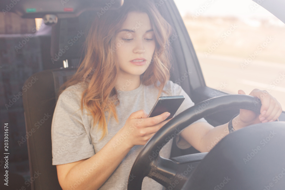 Lovely female driver holds smart phone, types text message on smart phone while stands on traffic light, poses in car, has concentrated look. Young woman driver uses online navigator for finding way