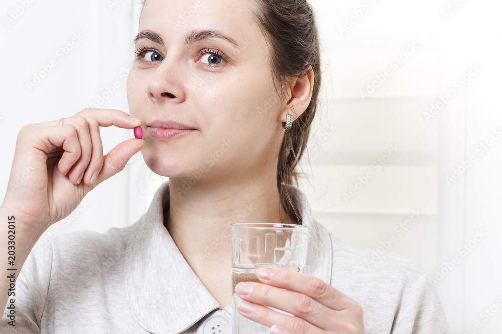 caucasian young brunette woman takes pill and drinks glass of clean water. girl is holding medicine and glass of water in hand. concept of health, therapeutic therapy. Medication. - obrazy, fototapety, plakaty 