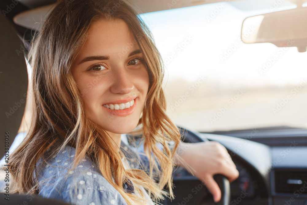 Back view of pretty smiling woman with broad smile, has attractive look, sits at wheel in car, has break after long trip, looks directly into camera. Positive woman drives automobile, enjoys speed