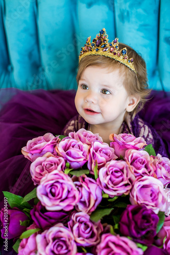Portrait of little princess baby in purple dress and diadem on blue sofa with bouquets of roses