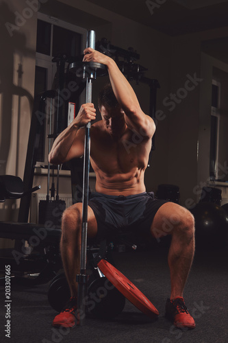 Muscular shirtless athlete holds barbell while sits on a bench in the gym.