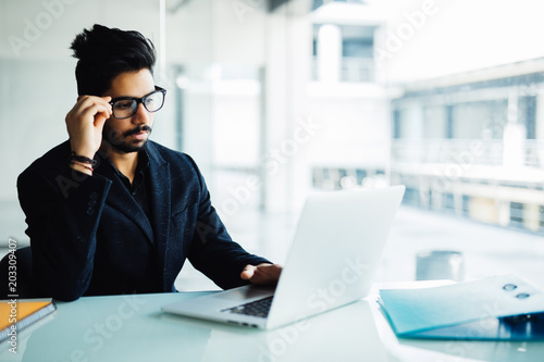Confused indian businessman working on laptop in office. Young indian man thinking over project at his desk with laptop.