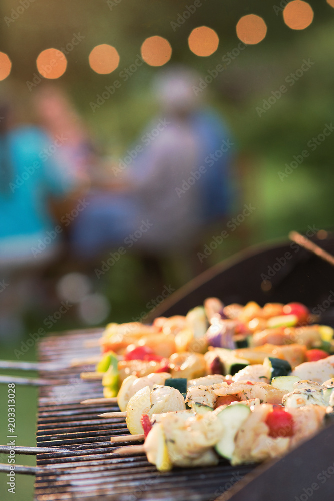 Fototapeta premium close-up of a barbecue where chicken skewers are cooking, friends gathered around a table to share a meal.