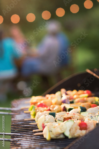 close-up of a barbecue where chicken skewers are cooking  friends gathered around a table to share a meal.