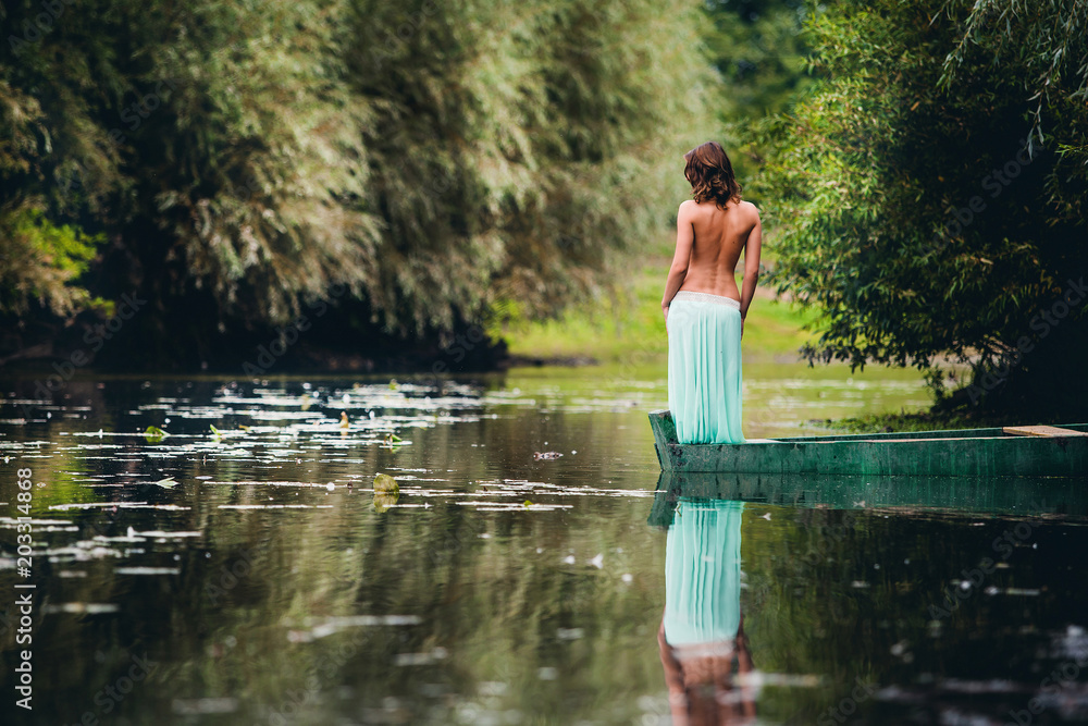 Fototapeta premium brunette in a turquoise skirt stands with a naked back in a boat on a river in the park in the summer