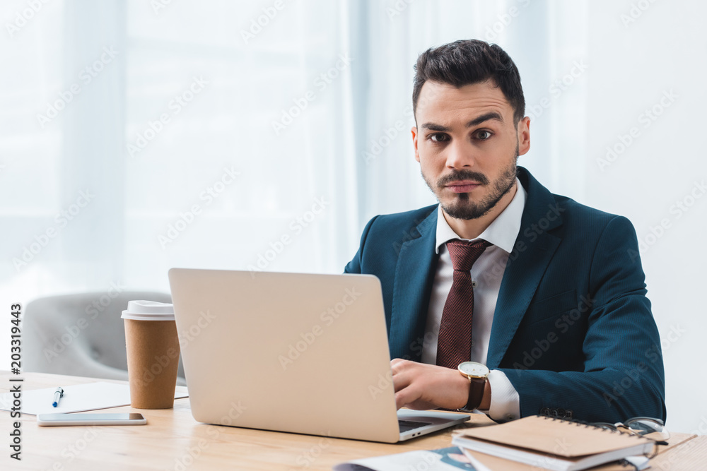 handsome young businessman using laptop and looking at camera in office handsome young businessman using laptop and looking at camera in office