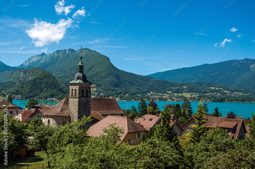 View of houses and belfry with blue sky mountains landscape on background, in the village of Talloires. A lovely village next to the Lake of Annecy. Department of Haute-Savoie, southeastern France.