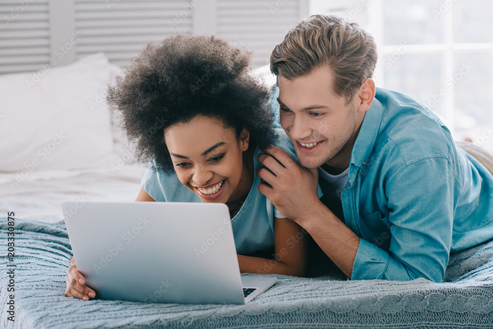 Multiracial couple lying on bed and looking at laptop screen
