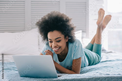 Smiling african american girl lying on bed and looking at laptop screen photo