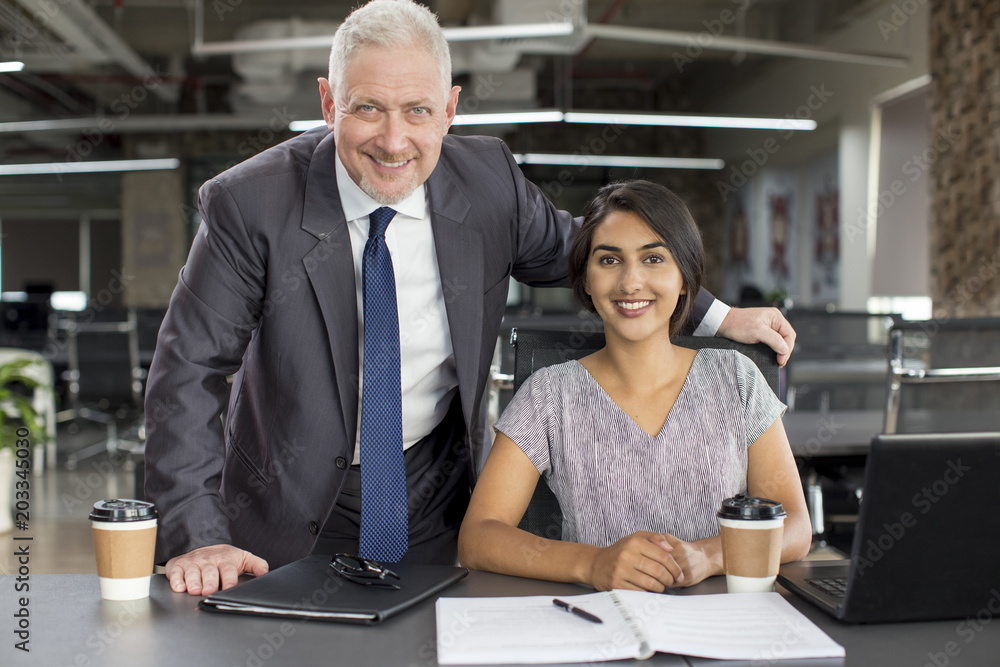 Business portrait of smiling experienced mentor and new hire. Positive mature man in suit posing with young Indian woman sitting at workplace. Mentorship and corporate portrait concept Business portrait of smiling experienced mentor and new hire. Positive mature man in suit posing with young Indian woman sitting at workplace. Mentorship and corporate portrait concept