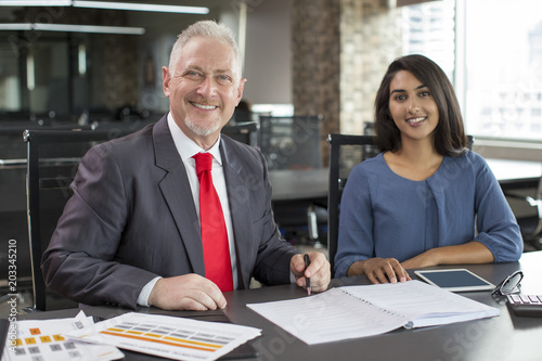 Portrait of smiling experienced mentor and young female employee. Mid adult man in formal jacket and Indian woman in casual wear posing at workplace. Business mentoring and teamwork concept