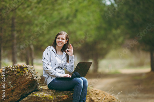 Young successful smart business woman or student in casual clothes sitting on stone using laptop and showing Ok sign in city park or forest working outdoors on green background. Mobile Office concept.