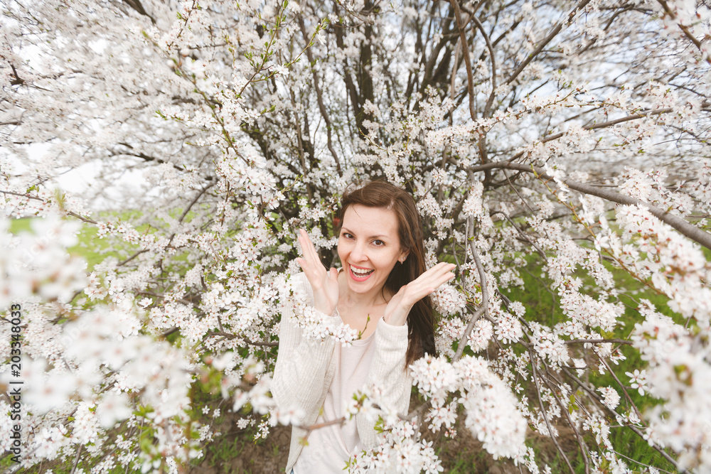 Young emotional smiling beautiful woman in light casual clothes keeping hands near face standing in city garden or park on blooming tree background. Spring nature, flowers. Lifestyle, leisure concept. Young emotional smiling beautiful woman in light casual clothes keeping hands near face standing in city garden or park on blooming tree background. Spring nature, flowers. Lifestyle, leisure concept.