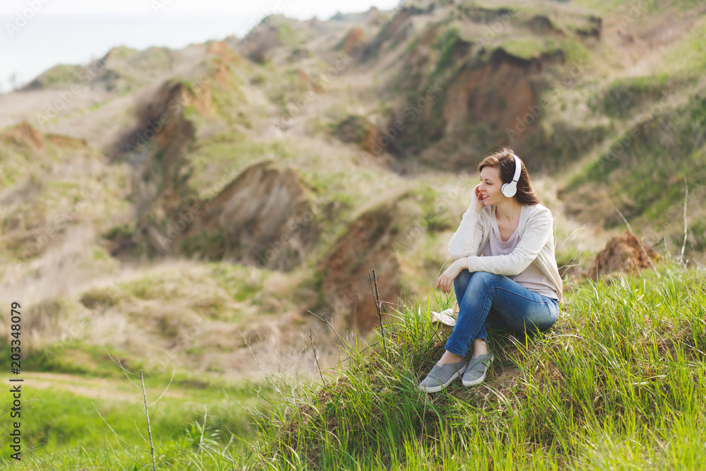 Young pensive relaxed beautiful woman in light casual clothes with headphones listening music sitting on grass with crossed legs looking aside on green field background. Lifestyle, leisure concept. Young pensive relaxed beautiful woman in light casual clothes with headphones listening music sitting on grass with crossed legs looking aside on green field background. Lifestyle, leisure concept.