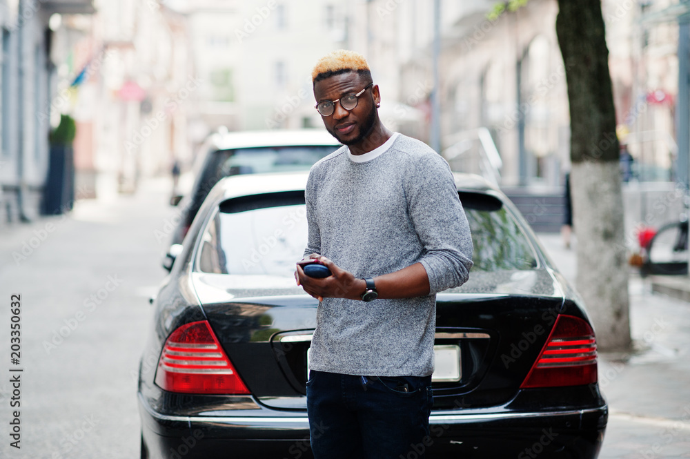 Fototapeta premium Stylish african american boy on gray sweater and glasses posed at street against black business car and speaking on phone. Fashionable black guy.