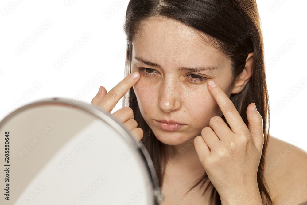 Beautiful young woman touch her face under eyes in mirror on white background Beautiful young woman touch her face under eyes in mirror on white background