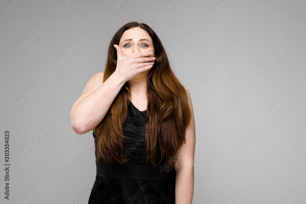 Emotional plus size model standing in studio closing mouth with hand showing surprise astonishment on gray background Emotional plus size model standing in studio closing mouth with hand showing surprise astonishment on gray background