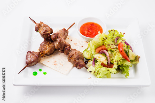 Shish kebab on bamboo skewers lies on a pita. Near sauce and fresh vegetable salad. White background (top view)