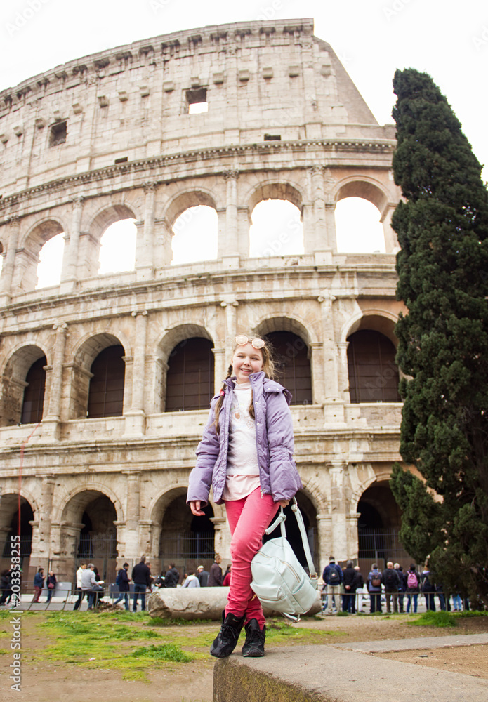 young little happy sunny girl in pink pants and glasses is standing fashion and posing on Colosseum background in Rome and smiling
