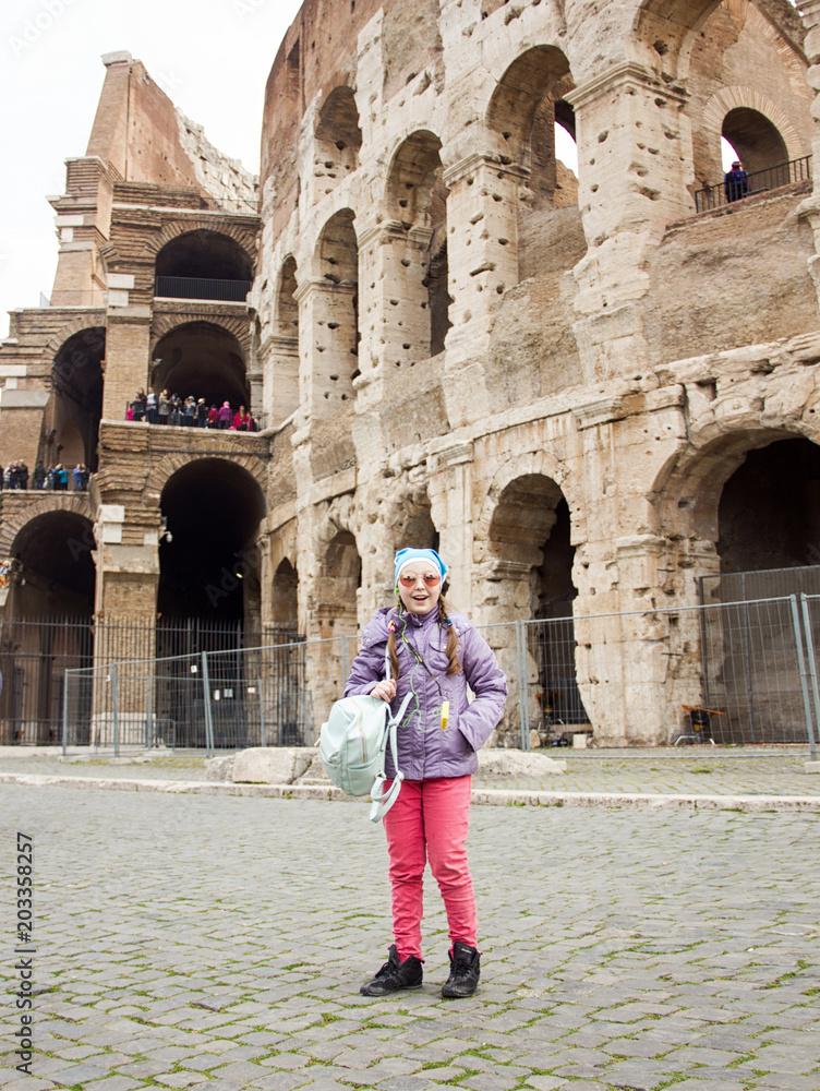 young little happy exciting girl in pink pants and glasses is standing fashion and posing on Colosseum background in Rome and smiling