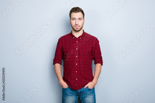 Serious confident concentrated strict successful guy clothed in red checkered shirt, holding hands in pockets of trousers, isolated on gray background, copy-space