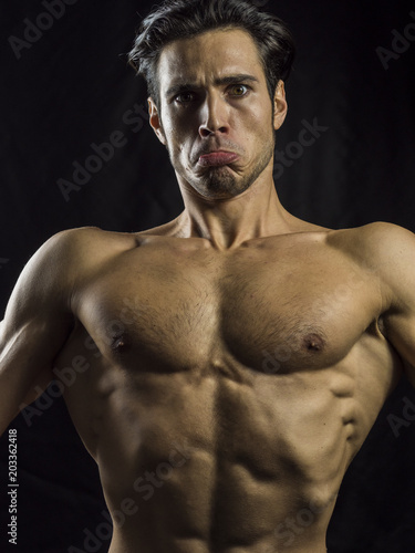 Young funny man showing muscular body with expression of effort looking at camera on black. 