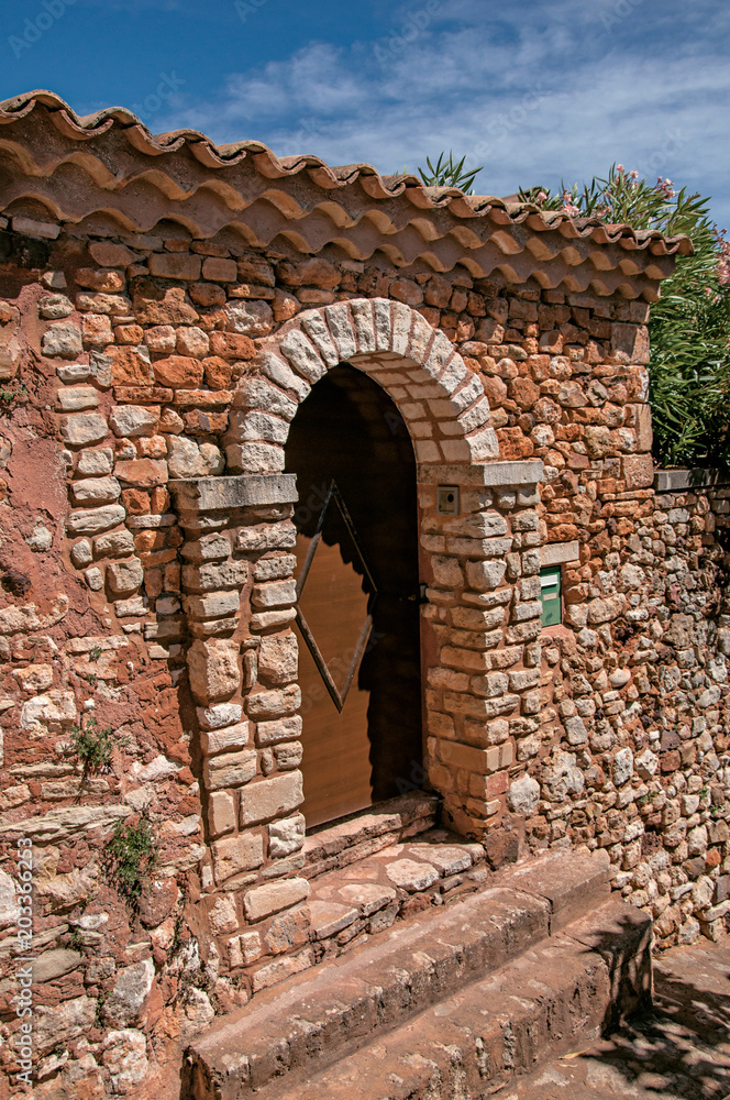 Naklejka premium View of wooden door and wall and arch in stone under a sunny blue sky, in the city center of the village of Roussillon. Located in the Vaucluse department, Provence region, southeastern France