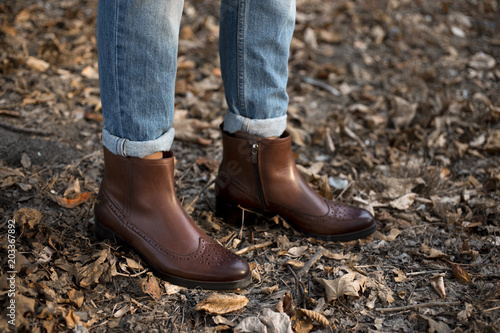 Brown shiny leather womens chelsea boots on woman legs with blue jeans in autumn forest or park.