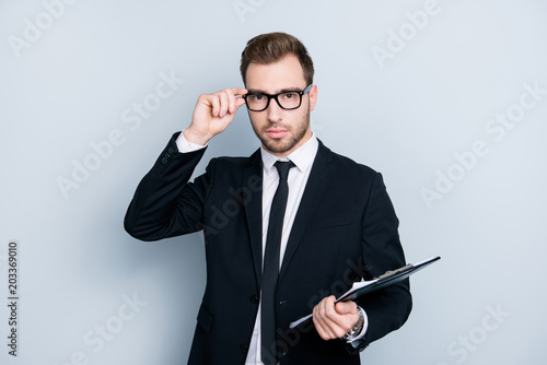 Portrait of attractive serious smart clever intelligent responsible punctual office worker wearing elegant suit holding clipboard touching glasses isolated on gray background