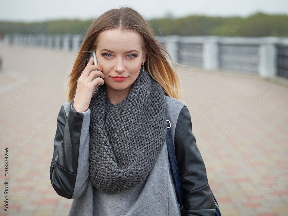 A stylish blonde in a fashionable urban style speaks on the phone, on the headset in a park on the river bank. The concept of spring mood