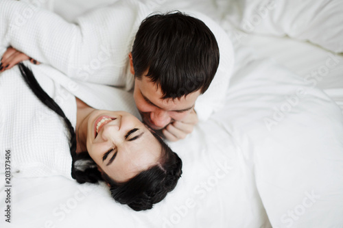 Young husband and wife of white coats play and smile in bed in a hotel room. Close up.