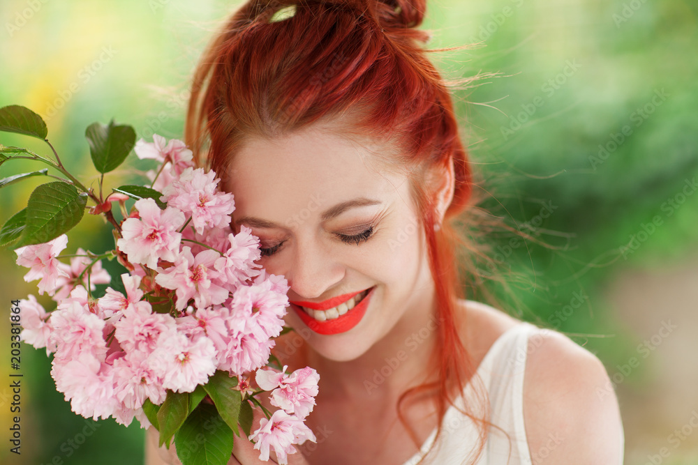 Beautiful young woman with red hair having fun standing in the garden with cherry blossom branch and perfect make up