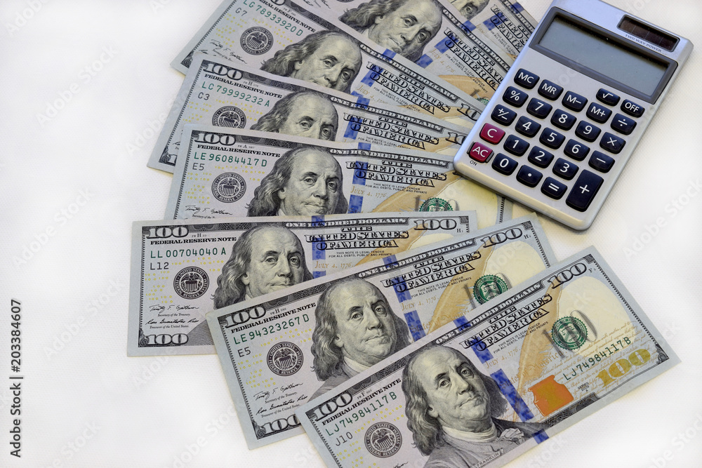 One hundred dollar bills spread out on table with calculator, white background. One hundred dollar bills spread out on table with calculator, white background.