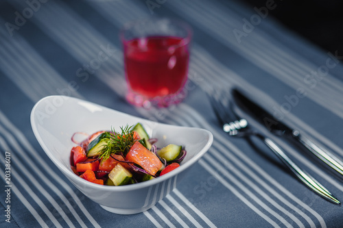 Table in the restaurant. On the table is a salad in a white plate  a knife  a fork  a glass with red juice. On the table is a blue tablecloth in strips.