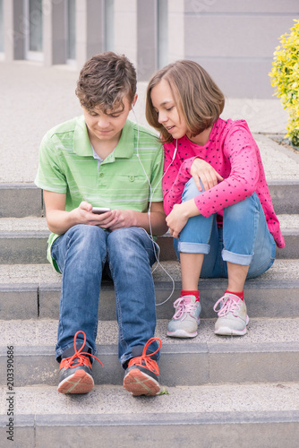 Happy teenage boy and girl with headphones are using gadget, talking and smiling while sitting on the stairs outdoors. Young sister and brother teens playing on mobile phone and listening to music. © valiza14
