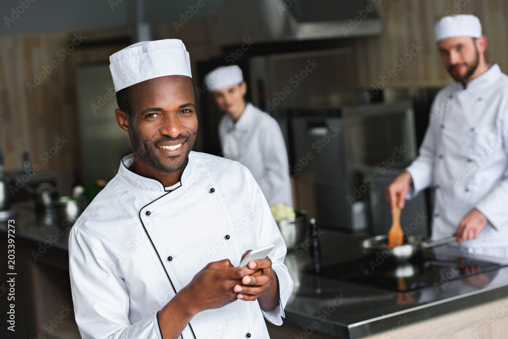 smiling african american chef using smartphone at restaurant kitchen and looking at camera