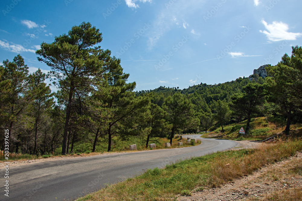 Road crossing forest and rock formations with blue and sunny sky, near the village of Baux-de-Provence. Bouches-du-Rhone department, Provence-Alpes-Côte d'Azur region, in southeastern France Road crossing forest and rock formations with blue and sunny sky, near the village of Baux-de-Provence. Bouches-du-Rhone department, Provence-Alpes-Côte d'Azur region, in southeastern France