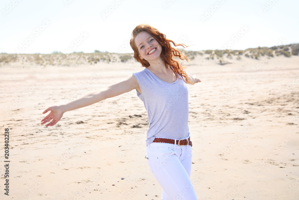 Hübsche rothaarige Frau steht mit ausgebreiteten Armen an einem Strand und lacht