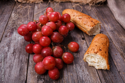 A bunch of juicy large red grapes and fresh crispy wheaten baguette on a table of dark old wooden boards