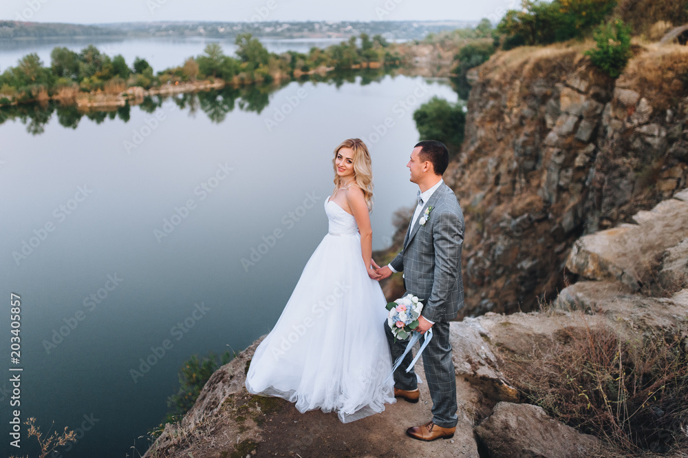 A pretty bride and a pretty bride with curly hair stand on a rug, holding hands, against the background of rocks and mountains. Portrait of a newlywed couple against the background of rocks.