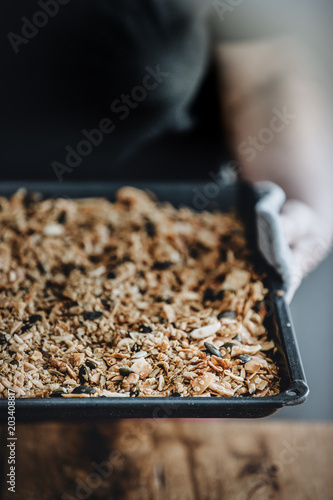 Woman holding a baking tray with freshly baked homemade granola. Healthy vegan snack easily made at home. Visible body parts of an elderly woman.