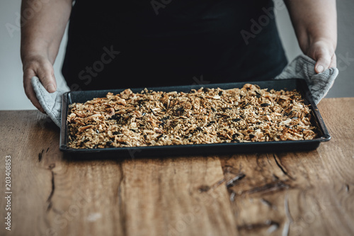 Woman holding a baking tray with freshly baked homemade granola. Healthy vegan snack easily made at home. Visible body parts of an elderly woman.