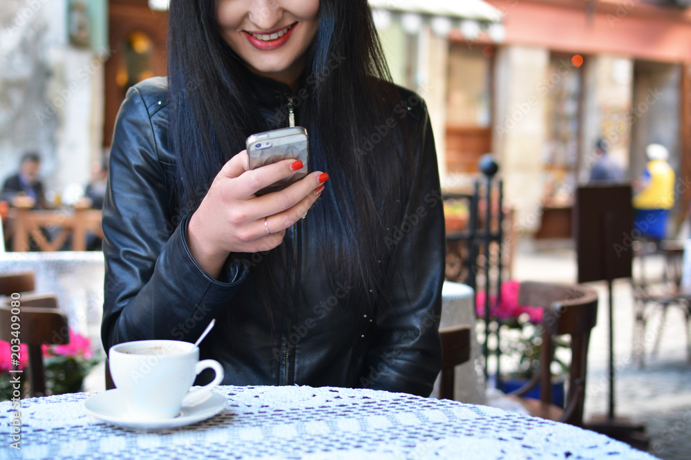 Attractive woman in a street cafe with a phone. A girl at a lunch break drinks coffee and communicates through the messenger with her smartphone. The Internet. Message. Attractive woman in a street cafe with a phone. A girl at a lunch break drinks coffee and communicates through the messenger with her smartphone. The Internet. Message.