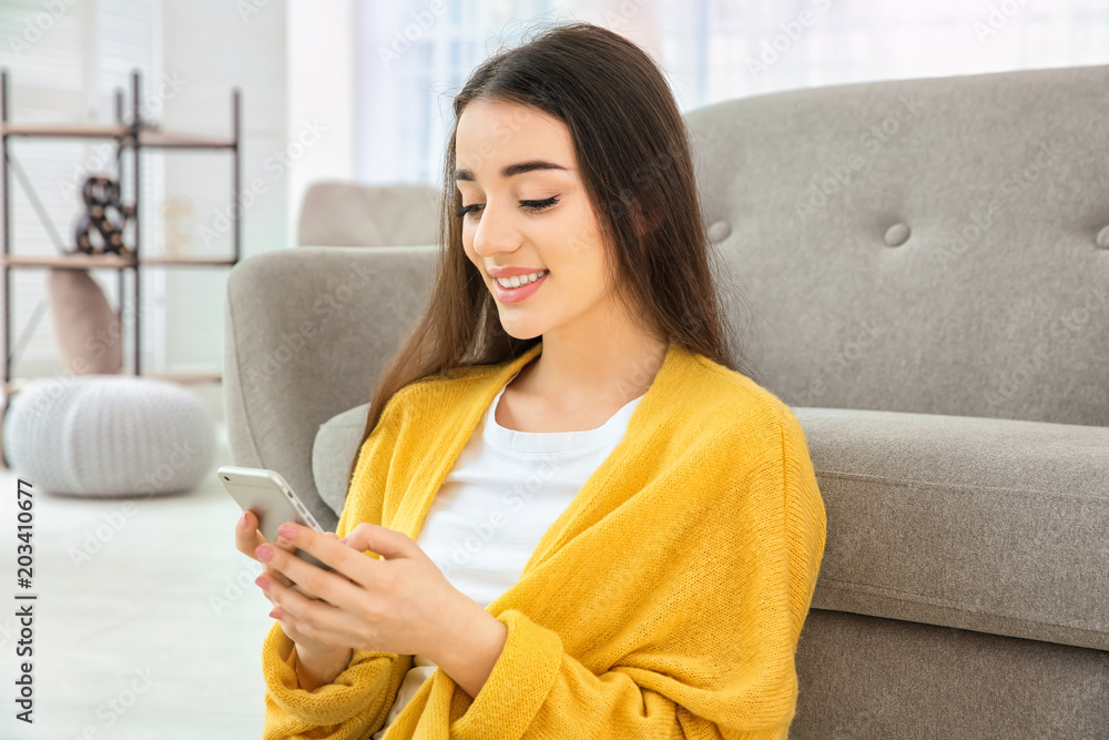 Attractive young woman using mobile phone sitting near sofa at home Attractive young woman using mobile phone sitting near sofa at home