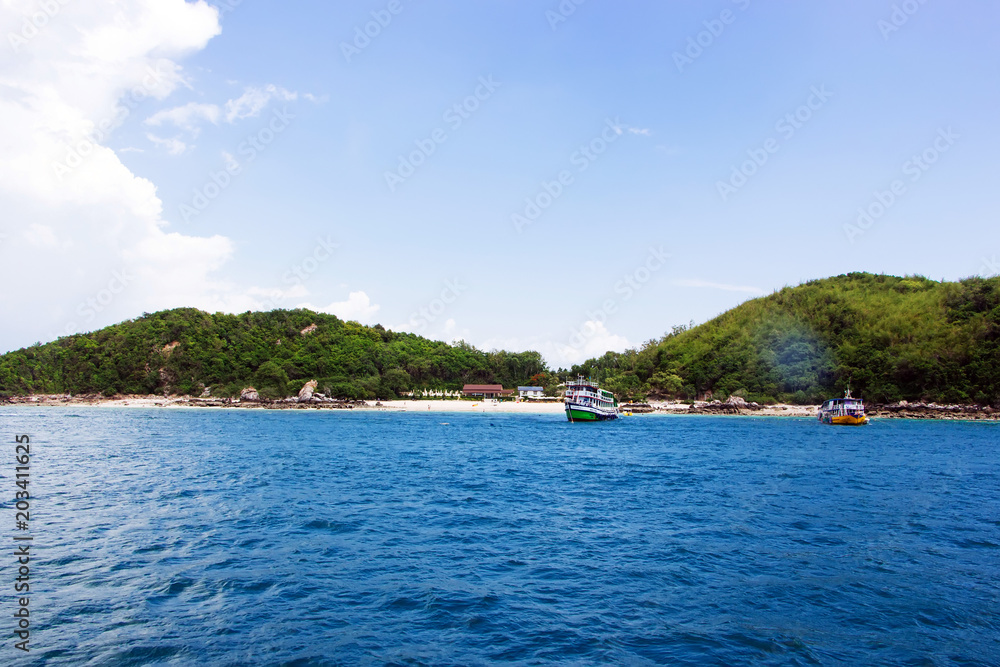 The beach on the island is surrounded by blue sea, light sky and foggy clouds. There is a boat and a resort on the island.