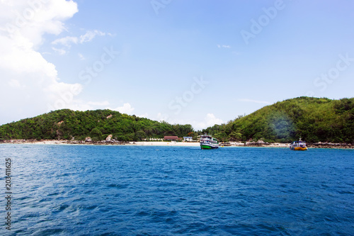 The beach on the island is surrounded by blue sea, light sky and foggy clouds. There is a boat and a resort on the island. © MPIX.TURE