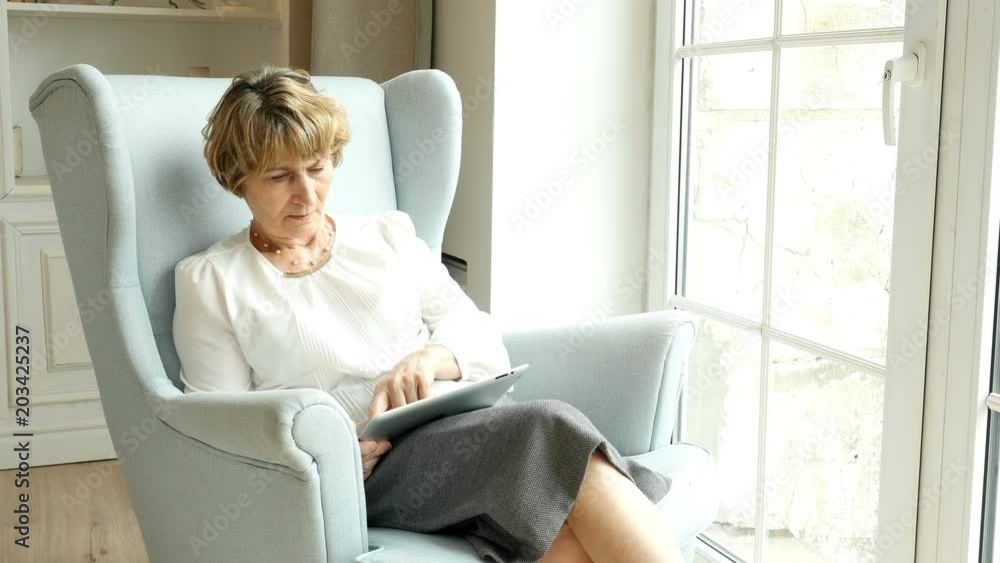 Senior attractive woman sits and websurfing on internet with tablet computer