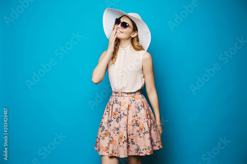Beautiful and fashionable blonde model girl in beige blouse, pink skirt, stylish sunglasses and hat smiling and posing in studio at blue background photo