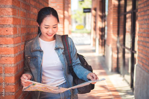 Happy beautiful young asian woman tourist with backpack on shoulders exploring map while standing in alley near vintage building, Holding map and street travel concepts.