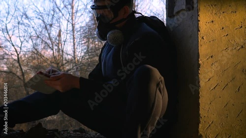 A young man sits in a ruined building in a respirator and glasses. He holds a tablet and a stylus. In the background trees. The concept of ecological disaster and post-apocalypse. photo